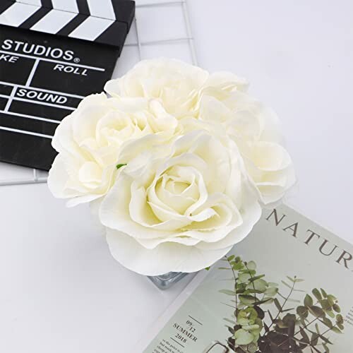 White roses in a vase on a table with a magazine and clapperboard.